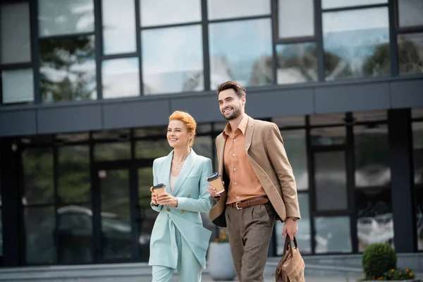 Успешные и стильные коллеги по бизнесу, гуляющие с кофе рядом с современным зданием на городской улице — стоковое фото
