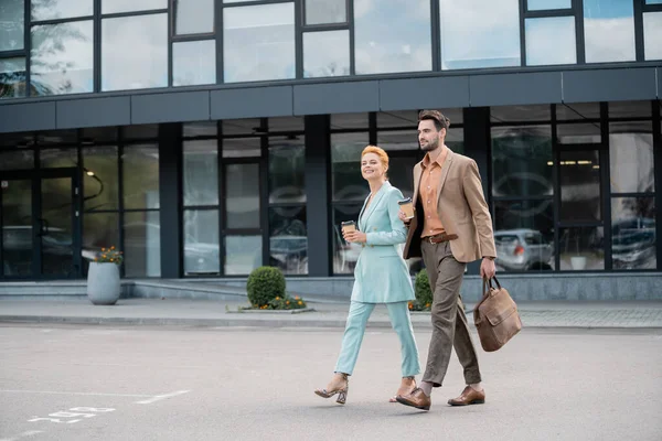 Full length of trendy business people walking with coffee to go near modern building on urban street — Stock Photo
