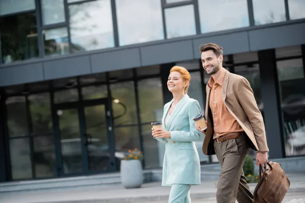 Glückliche und trendige Geschäftsleute, die mit Pappbechern in der Nähe eines modernen Gebäudes an der städtischen Straße spazieren gehen — Stockfoto