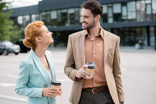 Trendy and pleased business partners with paper cups smiling at each other on urban street — Stock Photo