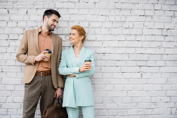 Pleased business colleagues in trendy suits holding paper cups and smiling at each other near brick wall — Stock Photo