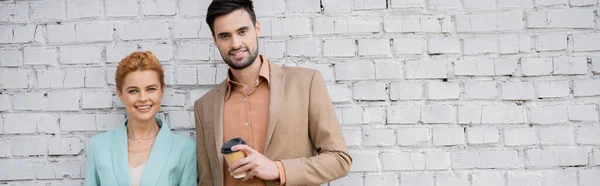 Homme d'affaires barbu avec café pour aller sourire à la caméra près de rousse femme d'affaires au mur de briques sur la rue, bannière — Photo de stock