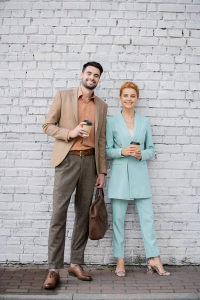 Cheerful business people with takeaway drinks looking at camera near brick wall on urban street — Stock Photo