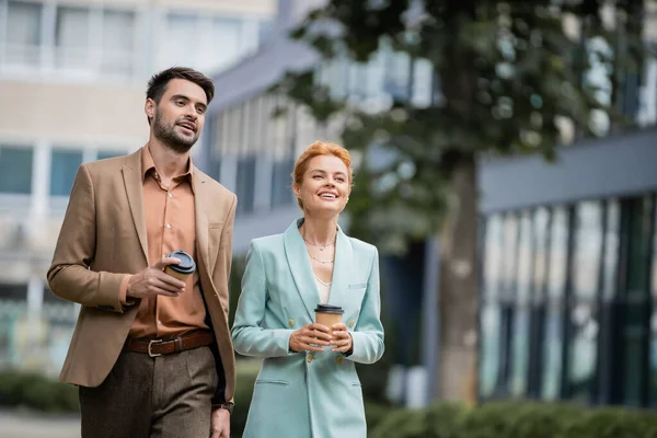 Веселые и стильные деловые коллеги с кофе идти улыбаясь и глядя в сторону на размытой городской улице — стоковое фото