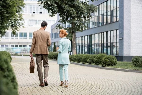 Visão traseira de mulher de negócios feliz e elegante falando com colega enquanto caminha ao longo de edifícios modernos na cidade — Fotografia de Stock