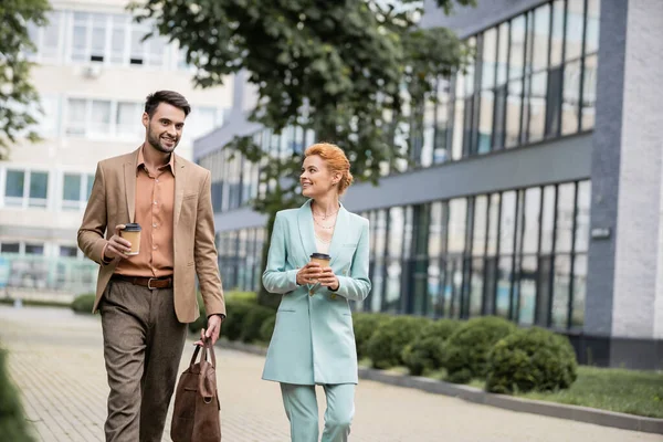 Pleased business partners in trendy suits holding paper cups and talking during walk in city — Stock Photo