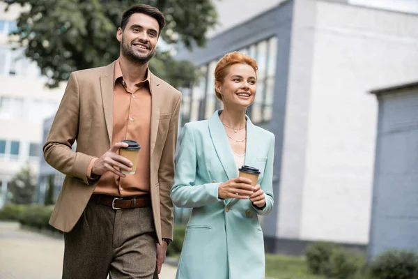 Joyful business partners in trendy suits walking with paper cups on street in city — Stock Photo