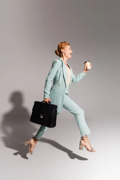 In voller Länge erstaunt Geschäftsfrau in stilvollem Anzug schwebt mit Coffee to go und schwarzer Aktentasche auf grauem Hintergrund — Stockfoto