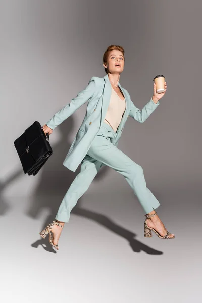 Piena lunghezza di donna d'affari eccitata in abito alla moda levitante con bevanda da asporto e valigetta in pelle su sfondo grigio — Foto stock