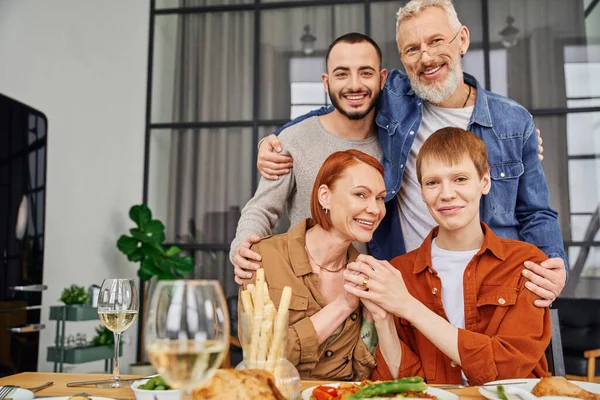 Glücklicher bärtiger Mann umarmt Sohn neben homosexuellem Partner am Tisch mit serviertem Abendessen — Stockfoto