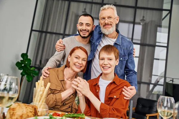 Jeune homme barbu avec des parents heureux et partenaire gay souriant à la caméra près de la table avec délicieux souper — Photo de stock