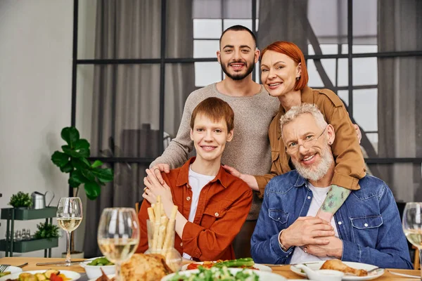 Joven y feliz pareja gay con sonrientes padres abrazando y mirando a la cámara cerca de la cena servida en sala de estar - foto de stock