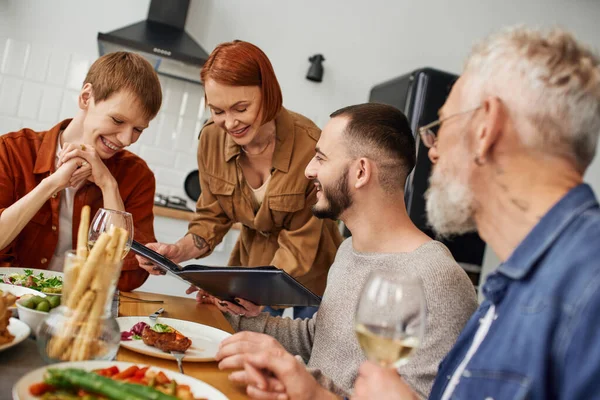 Smiling mother showing photo album to son next to his gay boyfriend during family supper at home — Stock Photo