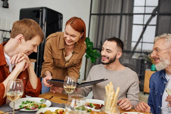 Happy redhead woman showing family photo album to son with gay boyfriend during supper at home — Stock Photo