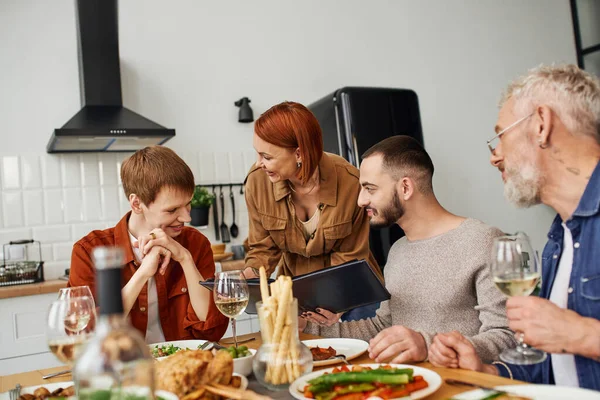 Cheerful redhead woman showing family photo album to smiling son with gay boyfriend near delicious supper served in kitchen — Stock Photo
