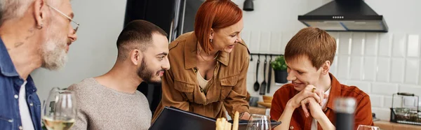 Рудий гей чоловік дивиться на фотоальбом біля хлопця і щасливої матері під час сімейної вечері вдома, банер — стокове фото