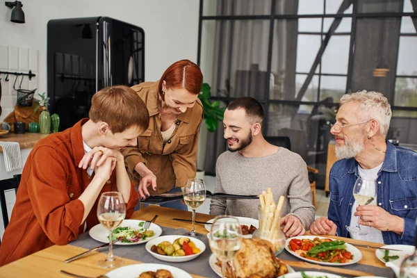 Redhead woman showing family photo album to son with gay boyfriend near delicious supper at home — Stock Photo