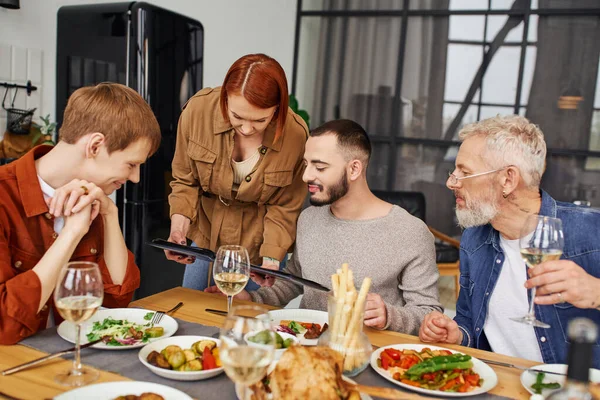 Молодые партнеры-геи смотрят фотоальбом во время вкусного семейного ужина на кухне — стоковое фото