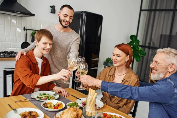 Feliz gay hombre tintineo vino vasos con novio y padres cerca delicioso cena en cocina - foto de stock