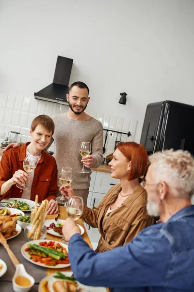 Jeune gay homme toasting avec vin pendant souper avec copain et heureux parents dans cuisine — Photo de stock