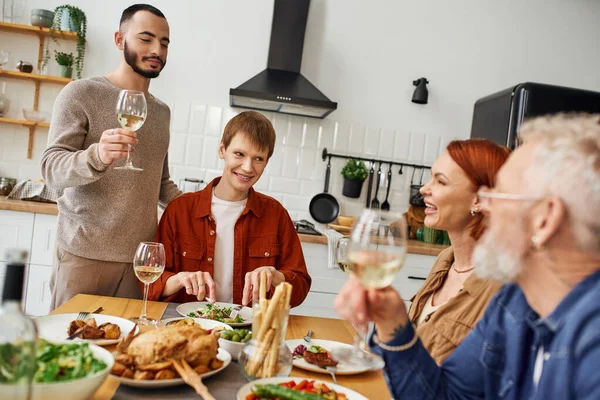 Barbudo gay hombre tostando con vino cerca novio y feliz padres durante familia cena en cocina - foto de stock