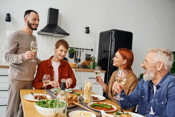 Feliz hombre tostando con vino durante la cena con la pareja gay y los padres en casa - foto de stock