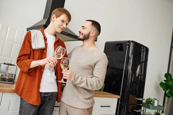 Happy gay couple clinking wine glasses and looking at each other in kitchen — Stock Photo