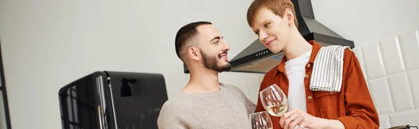 Young and happy gay couple clinking wine glasses and smiling at each other in kitchen, banner — Stock Photo