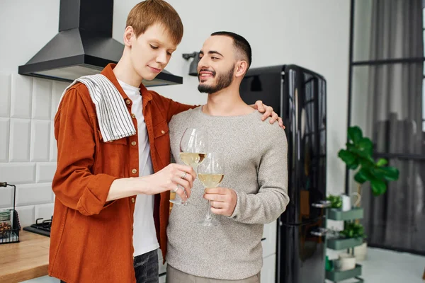 Redhead gay man with towel clinking wine glasses with bearded boyfriend in kitchen — Stock Photo