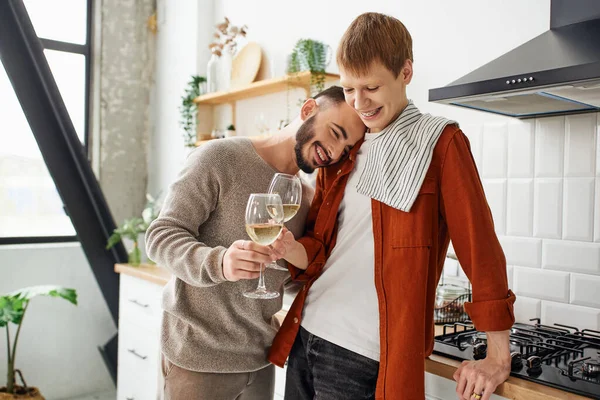 Alegre barbudo hombre apoyándose en pelirroja gay pareja y tintineo vino vasos en cocina - foto de stock