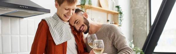 Задоволений бородатий чоловік, спираючись на рудого хлопця, стискаючи келихи для вина на кухні, банер — стокове фото