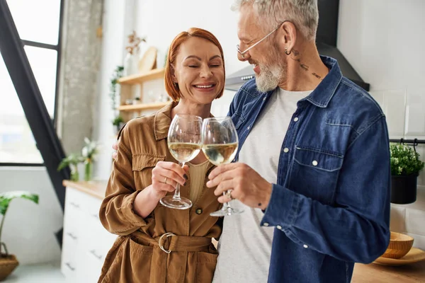 Весела і стильна пара середнього віку клонує келихи для вина на кухні — стокове фото