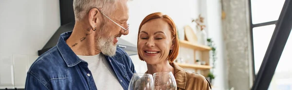 Bärtiger tätowierter Mann klimpert Weingläser mit fröhlicher rothaariger Frau in der Küche, Banner — Stock Photo
