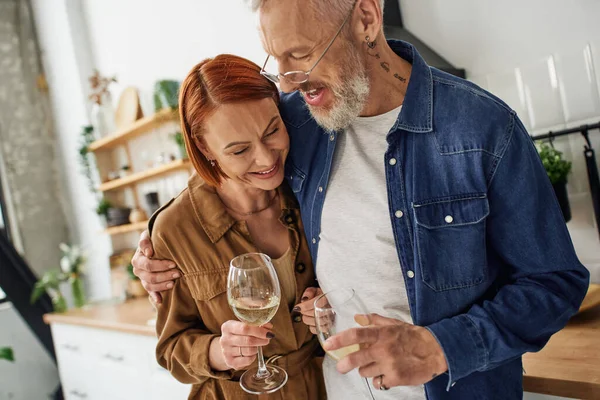 Cheerful bearded man embracing redhead wife while holding wine glasses in kitchen — Stock Photo