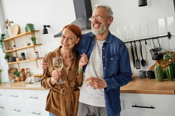Feliz pelirroja mujer sosteniendo copa de vino y sonriendo a la cámara cerca emocionado marido riendo en la cocina - foto de stock