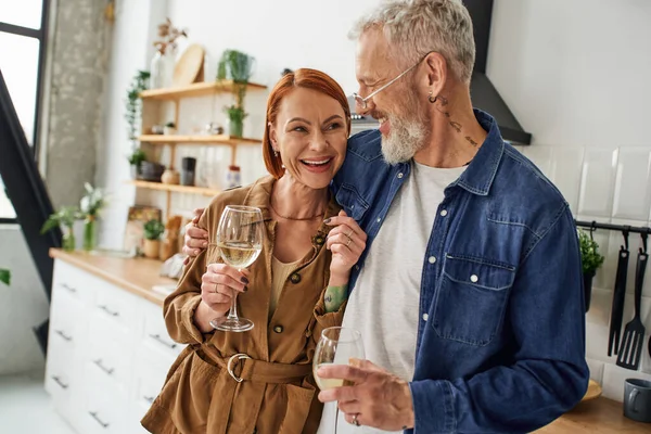 Bearded tattooed man embracing cheerful redhead wife while standing with wine glasses in kitchen — Stock Photo
