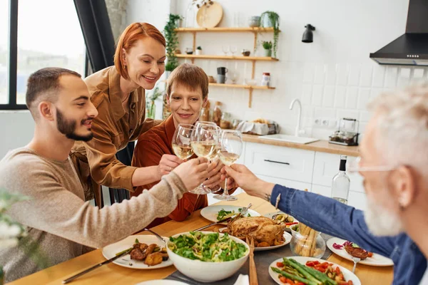 Sobregocijado gay hombre tintineo vino vasos con padres y novio cerca de familia cena en cocina - foto de stock