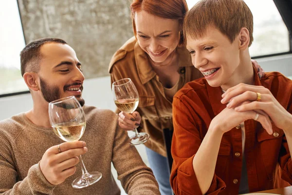 Cheerful redhead woman toasting with wine near young gay couple in kitchen — Stock Photo