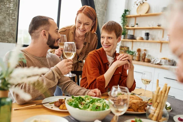 Souriant femme toasting avec vin près de fils avec gay copain près de délicieux repas dans cuisine — Photo de stock