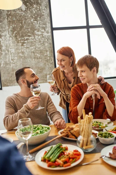 Збуджена руда жінка тости з вином біля щасливого сина з гей-партнером під час сімейної вечері на кухні — стокове фото