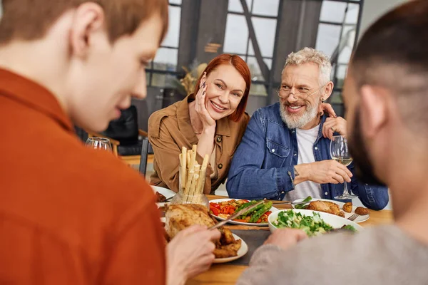Happy couple looking at son with gay partner having delicious supper in living room — Stock Photo