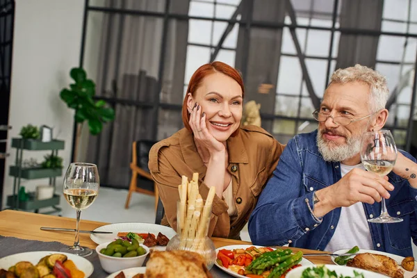 Happy redhead woman smiling near bearded husband with wine glass near delicious supper in living room — Stock Photo