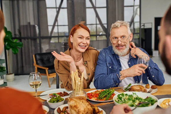 Задоволені батьки посміхаються біля розмитої гей-пари під час сімейної вечері у вітальні — стокове фото