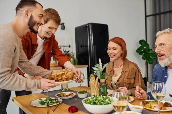 Joyful gay couple serving grilled chicken near happy parents in kitchen — Stock Photo