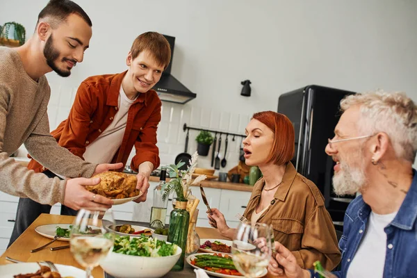 Веселые гей-партнеры держат жареного цыпленка рядом с родителями и стол подается с вкусным ужином на кухне — стоковое фото