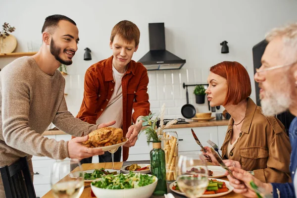 Allegra coppia gay che serve pollo alla griglia per cena in famiglia con i genitori in cucina — Foto stock