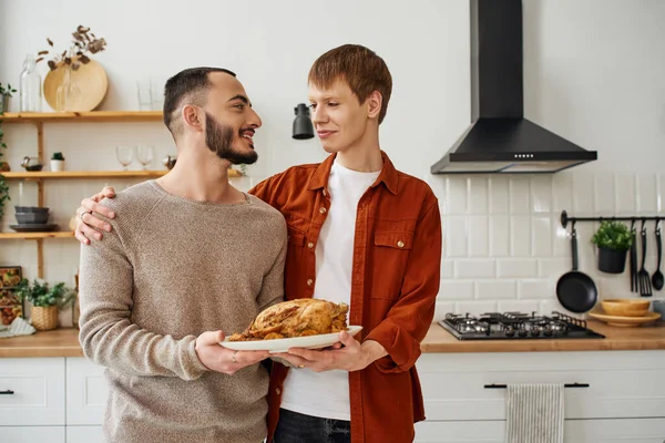 Complacida pareja gay sosteniendo delicioso pollo a la parrilla y sonriendo el uno al otro en la cocina - foto de stock
