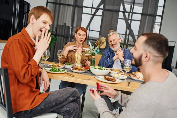 Расплывчатый гей с обручальным кольцом рядом с веселым парнем и изумленными родителями за ужином в гостиной — стоковое фото