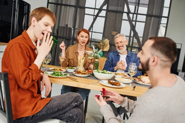 Изумленный гей, прикрывающий рот рукой рядом с бойфрендом и делающий предложение во время семейного ужина — стоковое фото