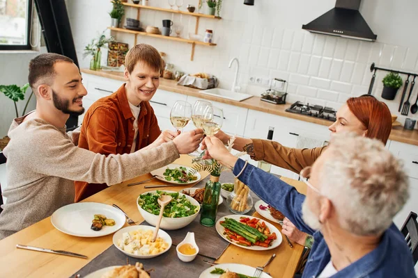 Allegro gay uomo clinking vino bicchieri con fidanzato e genitori vicino famiglia cena in moderno cucina — Stock Photo
